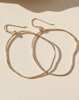 A gold large hoop earrings, each uniquely shaped, hang elegantly from simple hooks. Earrings are displayed on a cream backdrop.
