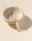 A gold hand-stamped, area code necklace with a delicate chain. Displayed on a small dish against a cream backdrop.