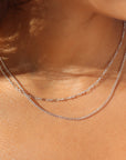 Extreme close-up shot of a woman's neckline highlighting two layered sterling silver chains in bright natural sunlight: a fine bar-and-link chain layered above a slightly longer delicate silver curb chain.