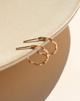 A wave like 14k gold fill textured huggie earring, displayed on a tan dish in the sunlight.