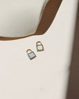 Gold padlock charm with shimmering white and blue opal stones on a neutral surface in warm natural light.