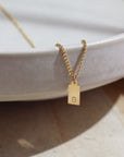 A gold rolo chain with a stamped mini tag with the letter "G". Necklace is displayed over a stone dish.