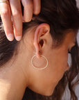 14k solid gold Organic Hoops by Token Jewelry. The hoops are a circle shape with hammered solid gold wire, photographed on a brunette model holding her hair back with one hand