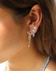 Side profile view of a woman with long dark hair displaying a beach-inspired silver ear stack: a dangling chain-link earring with a pearl drop in her main lobe, a seashell stud with tiny light-blue beaded drops in her second piercing, and a flat silver disk stud in her third, paired with a light-blue beaded necklace.