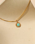 Gold necklace with a delicate box chain and a small round pendant featuring a bright turquoise stone set in a textured gold bezel, displayed on a sculptural cream ceramic stand in warm natural light.