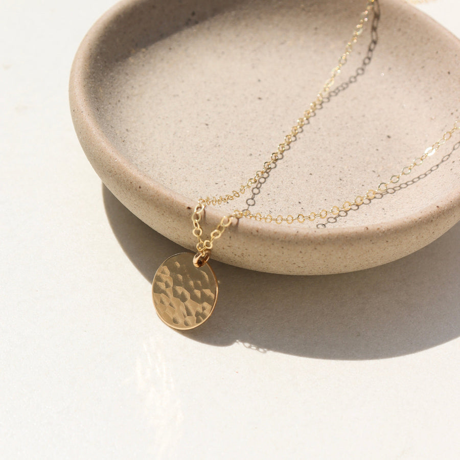 14k gold fill Moon drop necklace laid on a plate in the sunlight. This necklace features our simple chain met by a hammered coin, making it look like the moon.