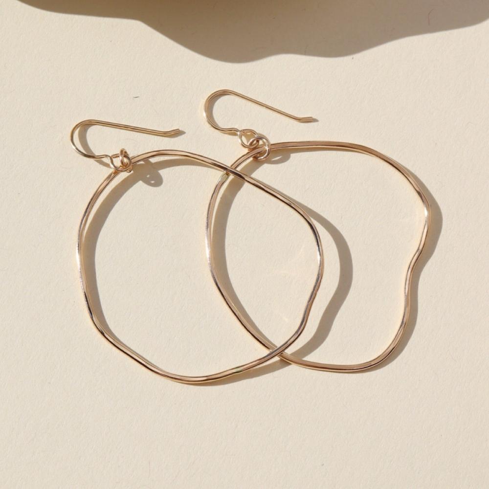 A gold large hoop earrings, each uniquely shaped, hang elegantly from simple hooks. Earrings are displayed on a cream backdrop. 