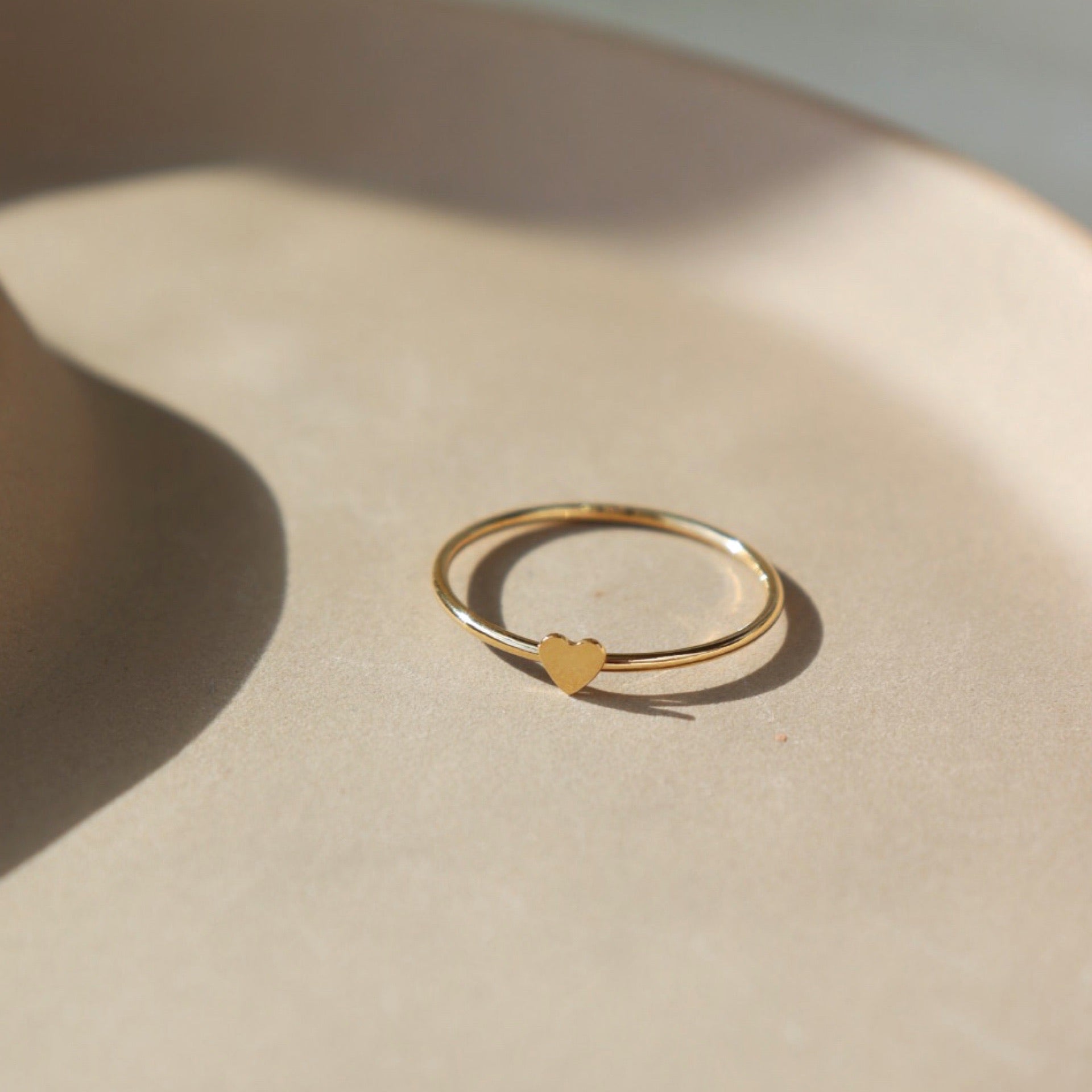 14k gold fill Tiny heart ring laid on a tan plate in the sunlight. This ring features a smooth band with a tiny heart. This ring is so cute stacked with any of the stacking rings at Token Jewelry. 