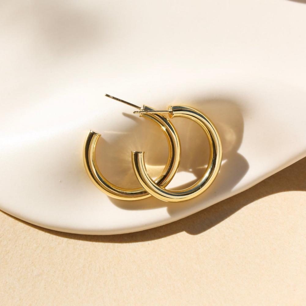 14k gold fill Harper Hoops laid on a tan paper in the sunlight. These earrings feature a classic smooth hoop with a post making the easy to take in and out.