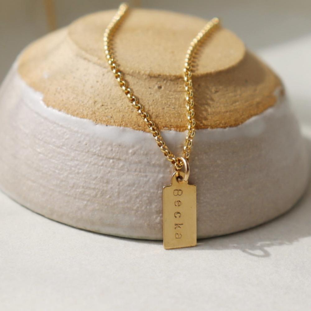 14k gold fill Mini Tag Personalized Necklace placed on a tan and gray bow. This necklace features the coin necklace chain followed by a mini tag with personalization of your choice.