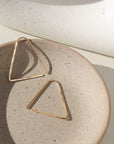 14k gold fill Sunday Earrings laid on a gray plate in the sunlight. These earrings feature a triangle look easy to thread in your ear.