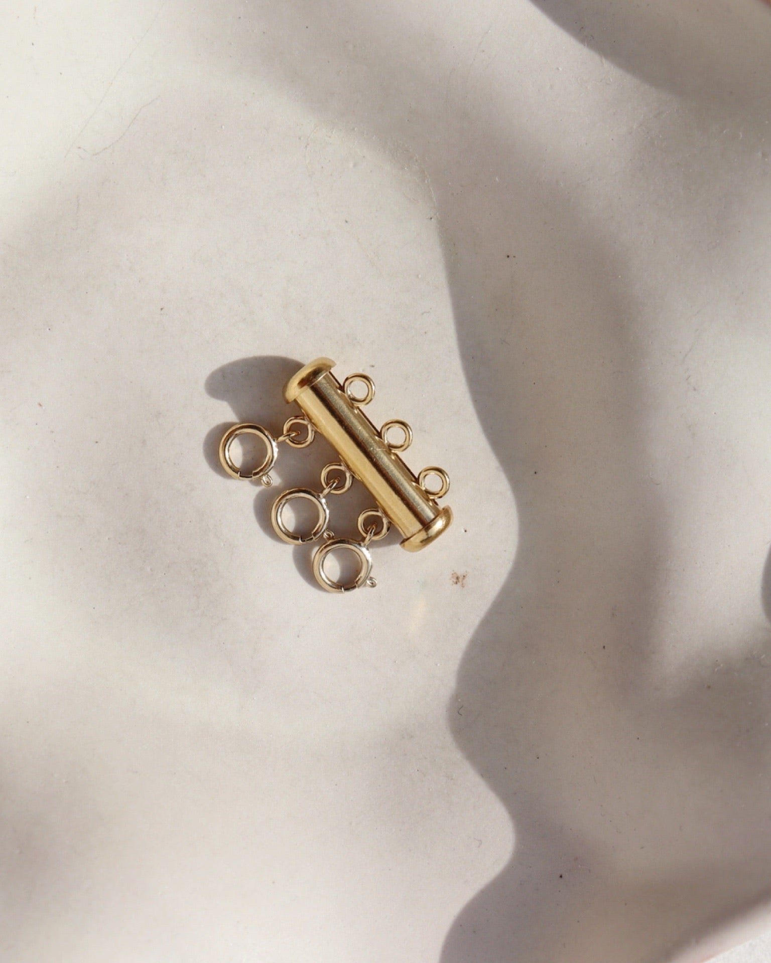 A gold necklace layering clasp with three necklace attachment points laid on a cream backdrop. 
