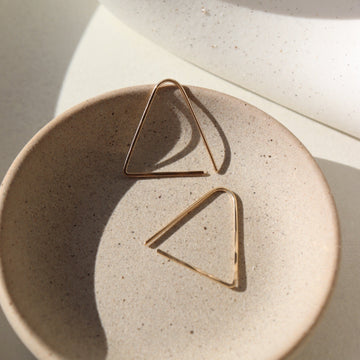 14k gold fill Sunday Earrings laid on a gray plate in the sunlight. These earrings feature a triangle look easy to thread in your ear.