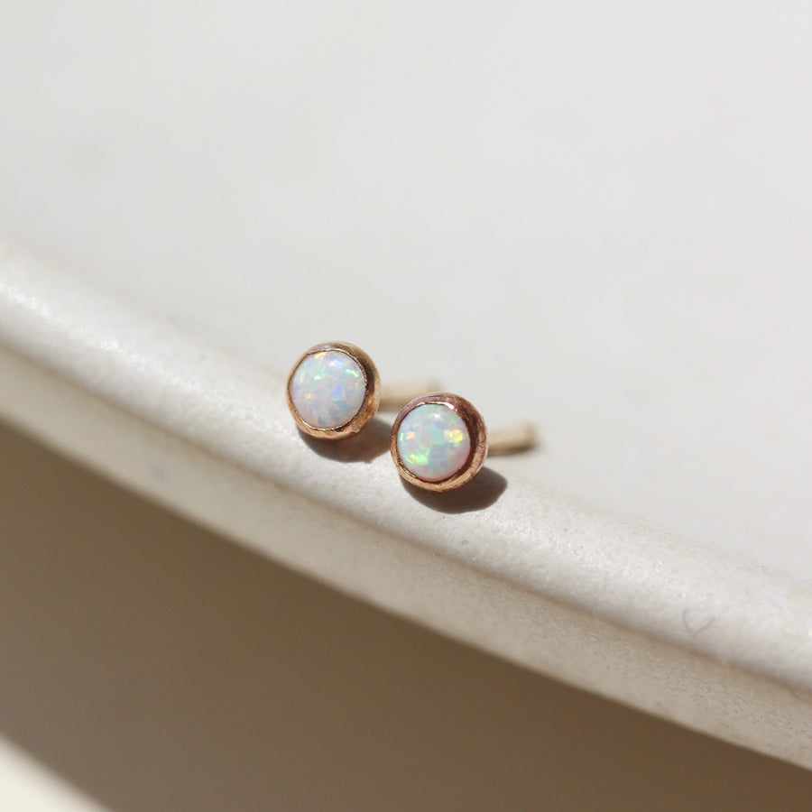 14k gold fill Petite opal studs laid on a tan plate in the sunlight. these earring feature a 3mm opal gemstone followed by a bezel ear post.