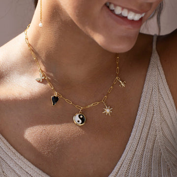 14k gold fill chain with various gold charms, featuring a Butterfly, black heart, yin yang symbol, opal star, and a fleur de lis, photographed on a model wearing a beige knit tank top
