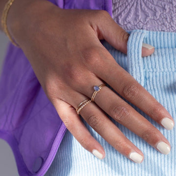 Model wearing 14k gold fill Tanzanite Ring. This ring features a simple smooth band featuring a Tanzanite cab. The model is also wearing other 14k gold fill bands, light blue corduroy shorts, a lavender purple textured top, and a purple vest
