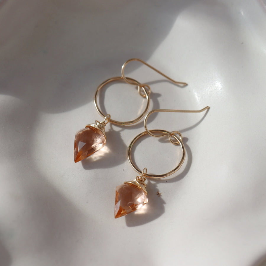 Champagne Quartz Drops laid on a white colored plate laid in the sunlight. These earrings feature a hook earring with a open circle disc followed my the champagne quartz gemstone.