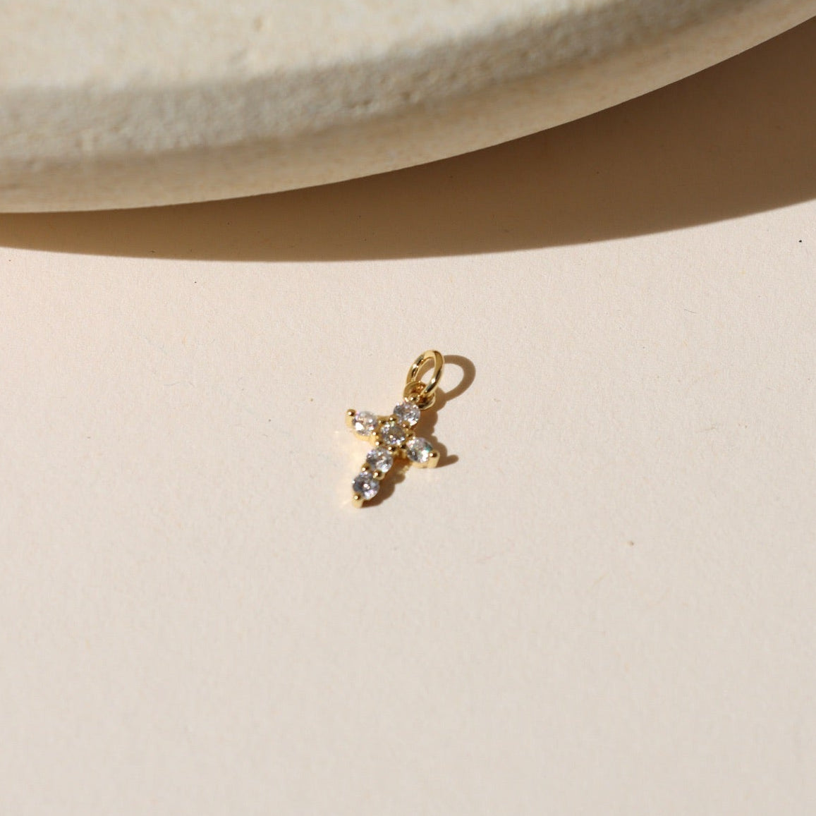 Gold cross charm with clear sparkling stones, photographed on a neutral surface in warm natural light.