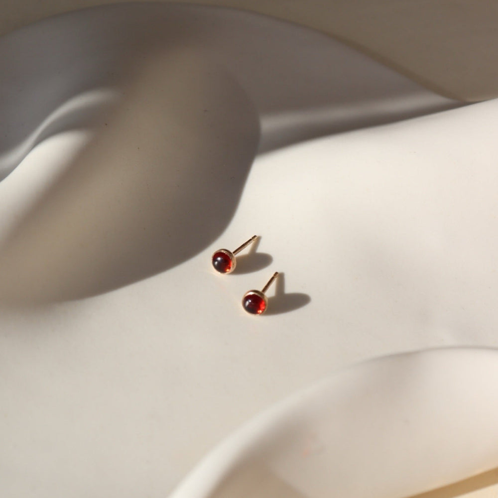 4mm Red Garnet Stud Earrings with 14k gold fill post and bezel, photographed on a curvy white ceramic dish