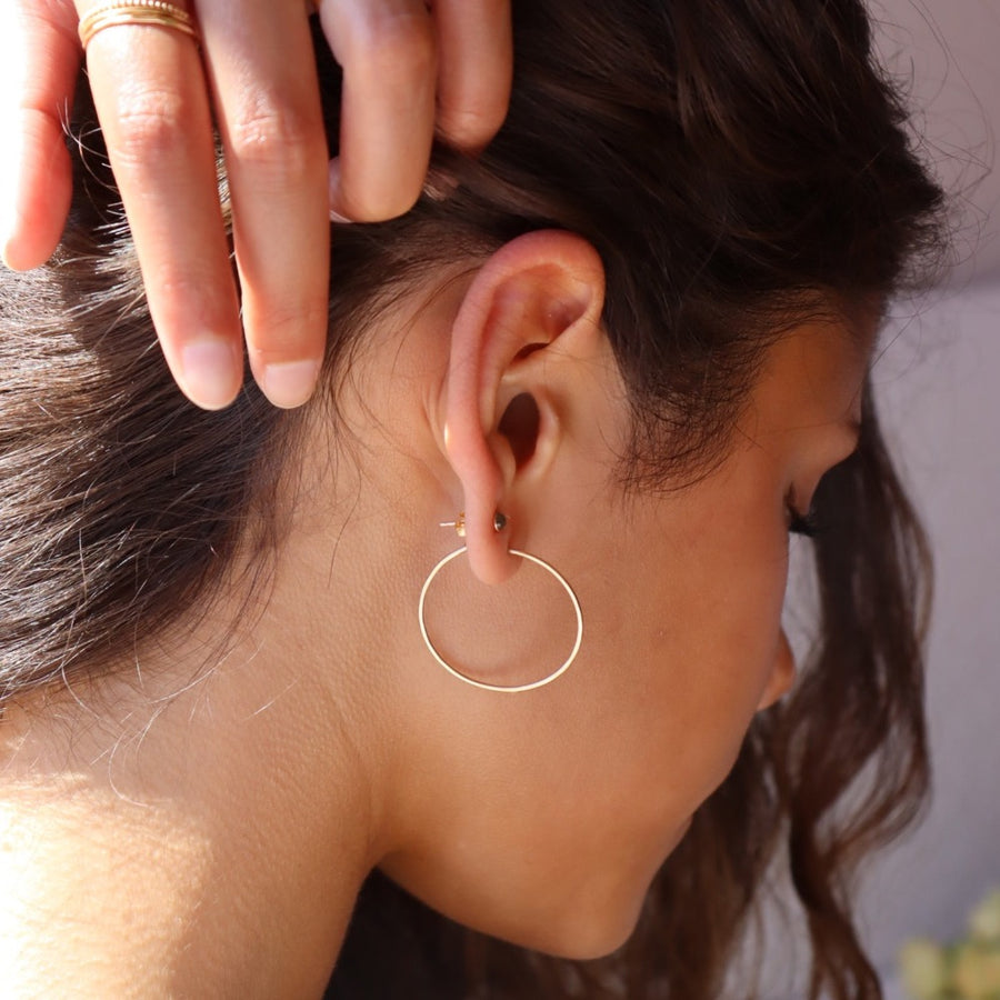 14k solid gold Organic Hoops by Token Jewelry. The hoops are a circle shape with hammered solid gold wire,  photographed on a brunette model holding her hair back with one hand