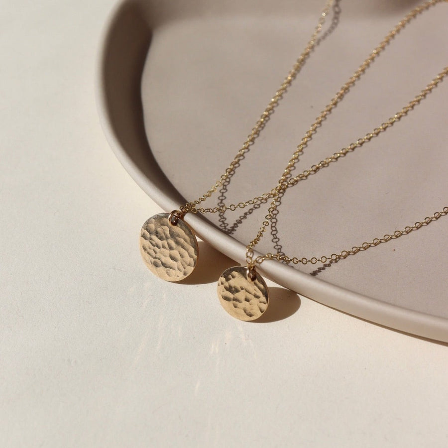 14k gold fill sun drop necklace laid on a tan plate in the sunlight. This necklace features our simple chain met by a hammered coin, making it look like the moon.