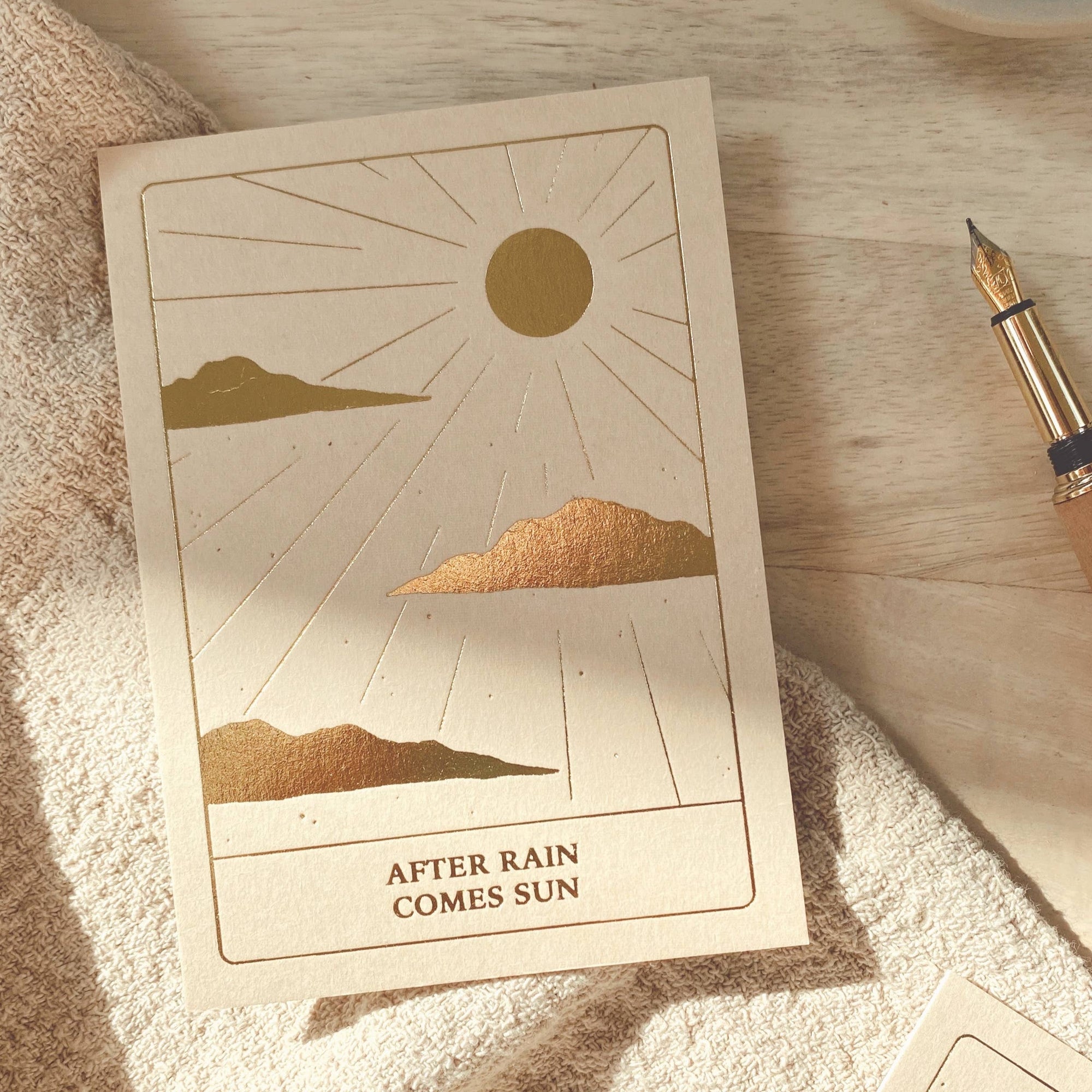 A card with a sun shining along with cloud on the bottom of the card it says &quot;after the rain comes the sun.&quot; That is set on a blanket layer across the table.