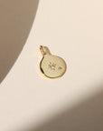 Gold coin charm with a starburst crystal detail and small accent stone, photographed on a warm neutral surface with soft natural light and shadows.