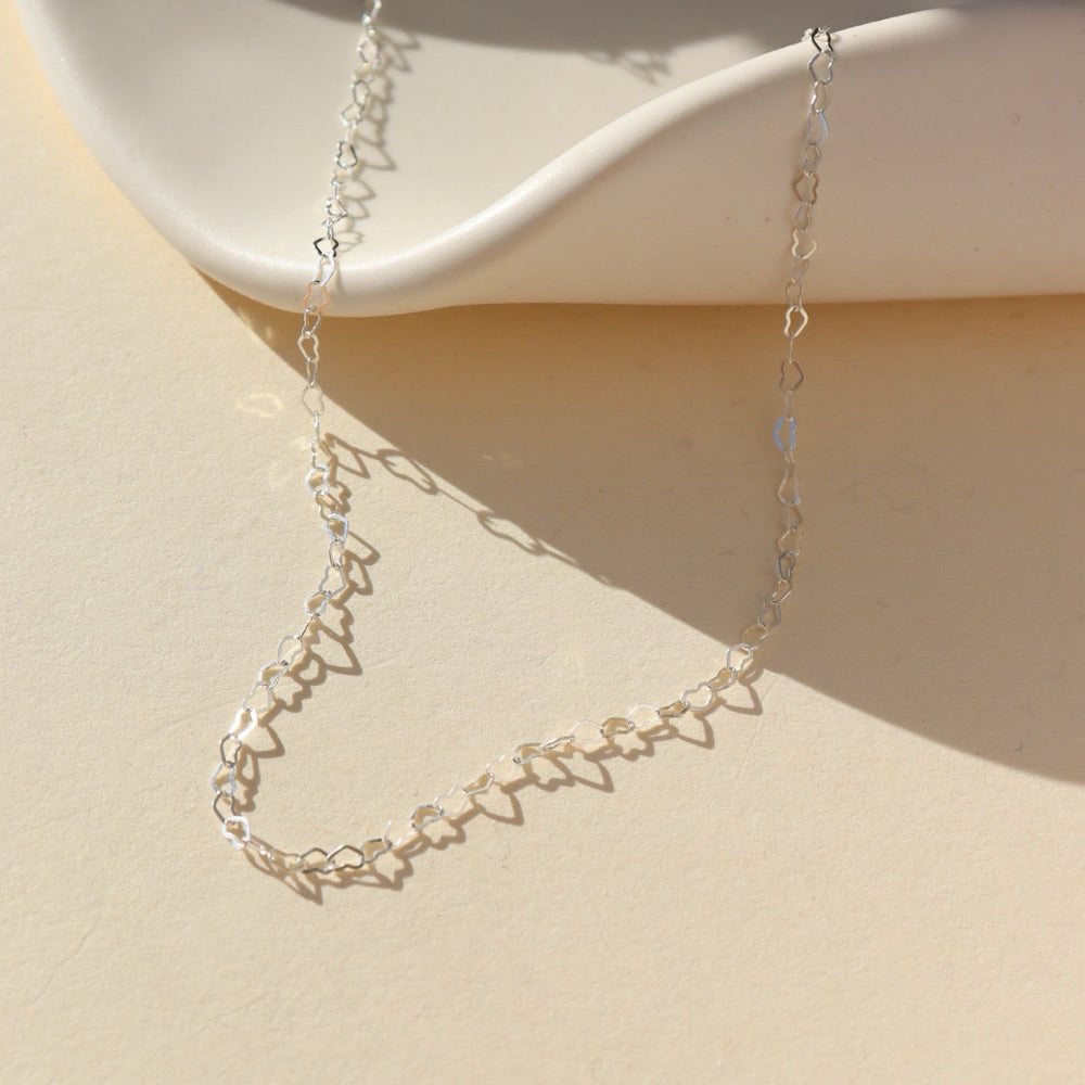 925 Sterling Silver Petite heart chain from Token Jewelry, photographed on a tan, organically shaped dish. This chain features a silver tiny heart wire throughout the whole chain. 