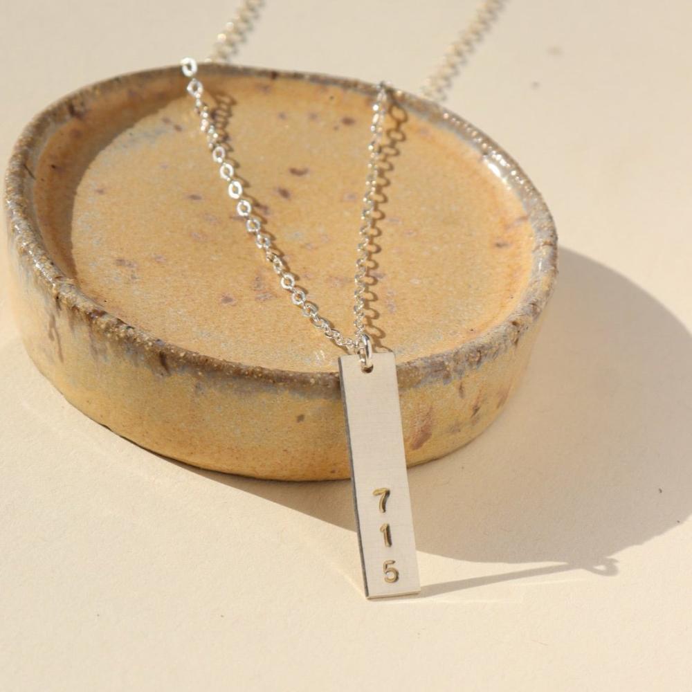 A silver hand-stamped, area code necklace with a delicate chain. Displayed on a small dish against a cream backdrop. 