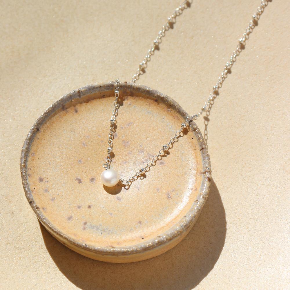 925 sterling silver beaded chain with a pretty single pearl necklace laid on a ceramic dish in the sunlight. 