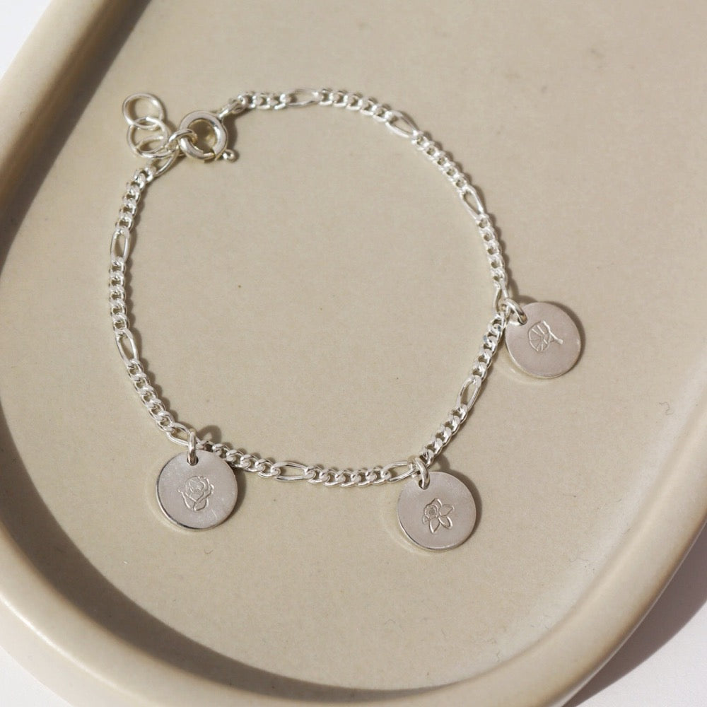 925 sterling silver Birth Flower Charm Bracelet laid on a tan plate in the sunlight. This Bracelet feature the Carter chain with as many birth flower charms as you want.