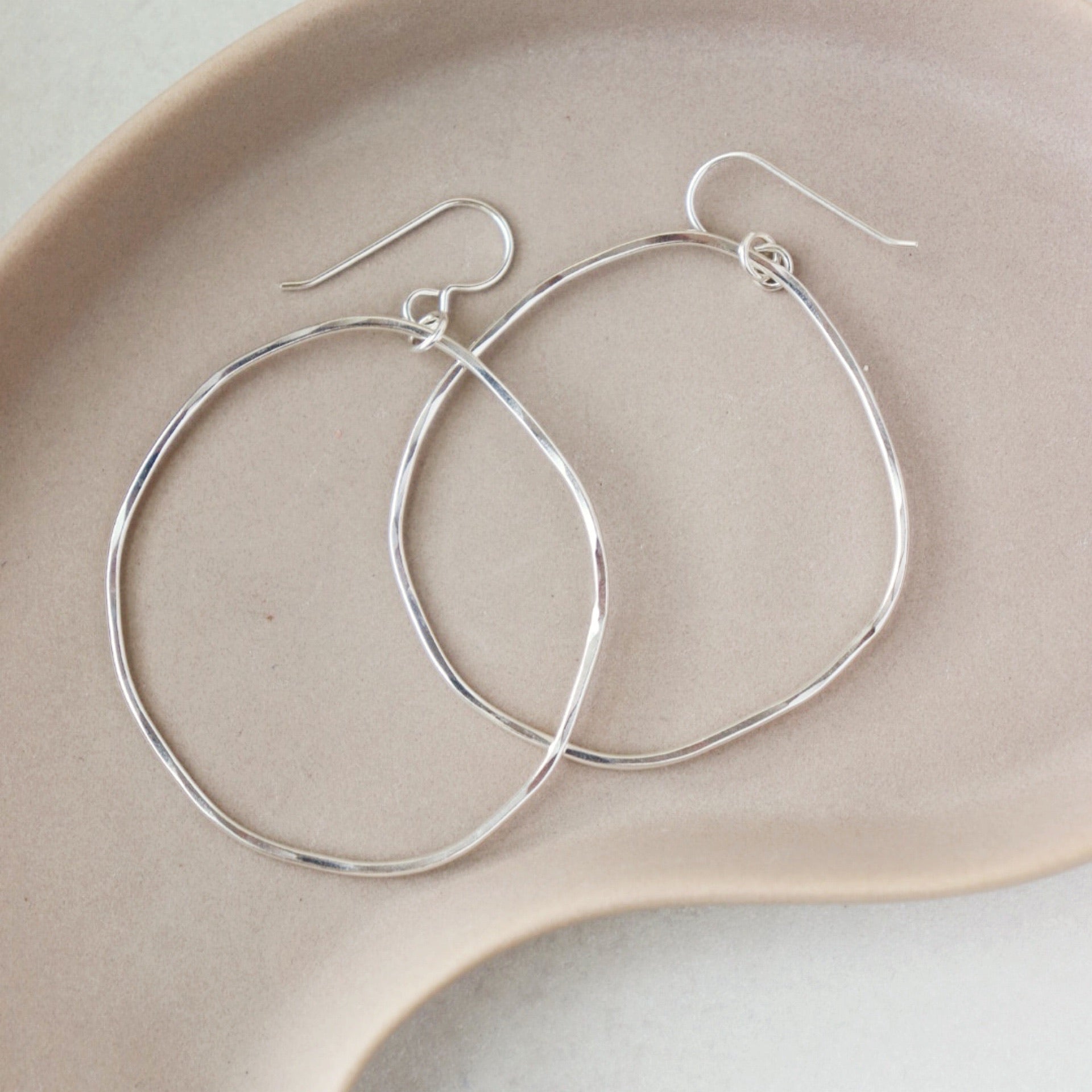 A silver large hoop earrings, each uniquely shaped, hang elegantly from simple hooks. Earrings are displayed on a dish. 