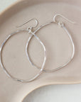 A silver large hoop earrings, each uniquely shaped, hang elegantly from simple hooks. Earrings are displayed on a dish. 