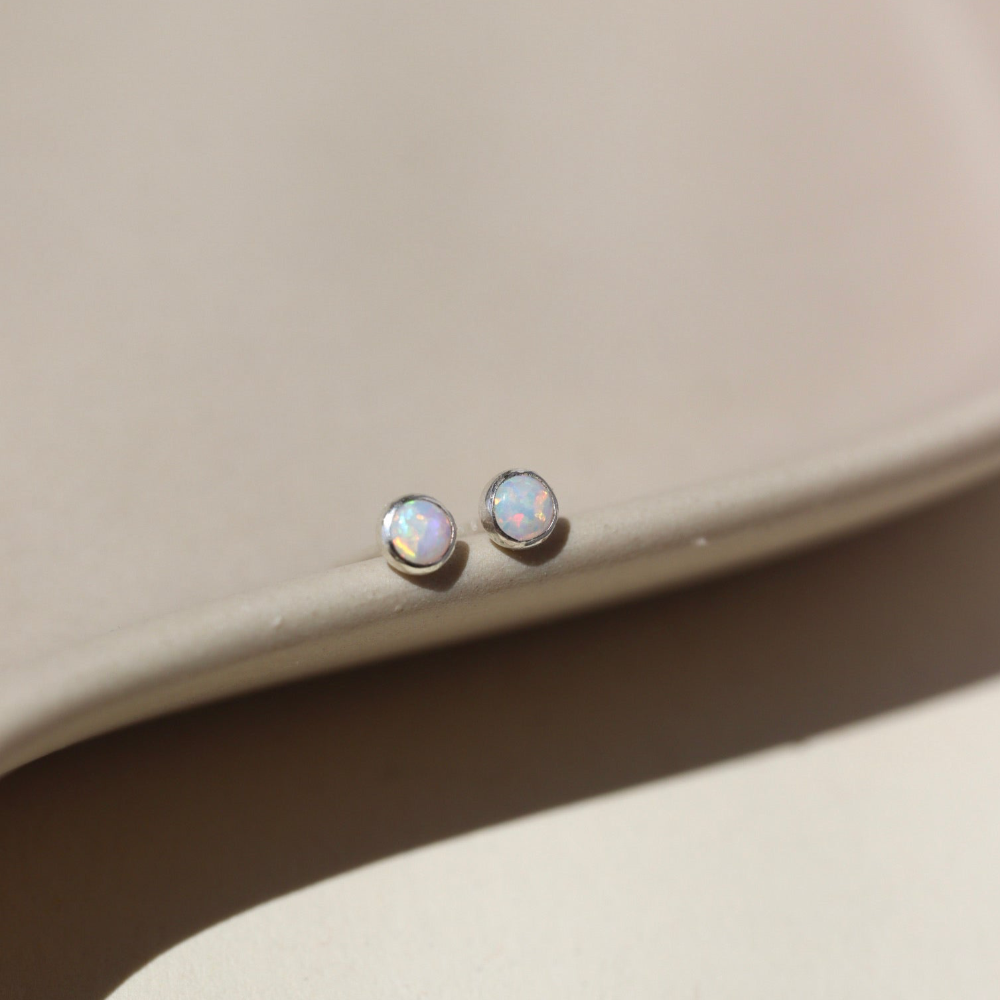 14k gold fill Petite opal studs laid on a tan plate in the sunlight. these earring feature a 3mm opal gemstone followed by a bezel ear post.