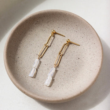 14k gold fill Lucy Drop earrings laid on a tan plate in the sunlight. These earrings feature a stud earring with a chain link dangle followed by two rainbow moonstone gemstones.