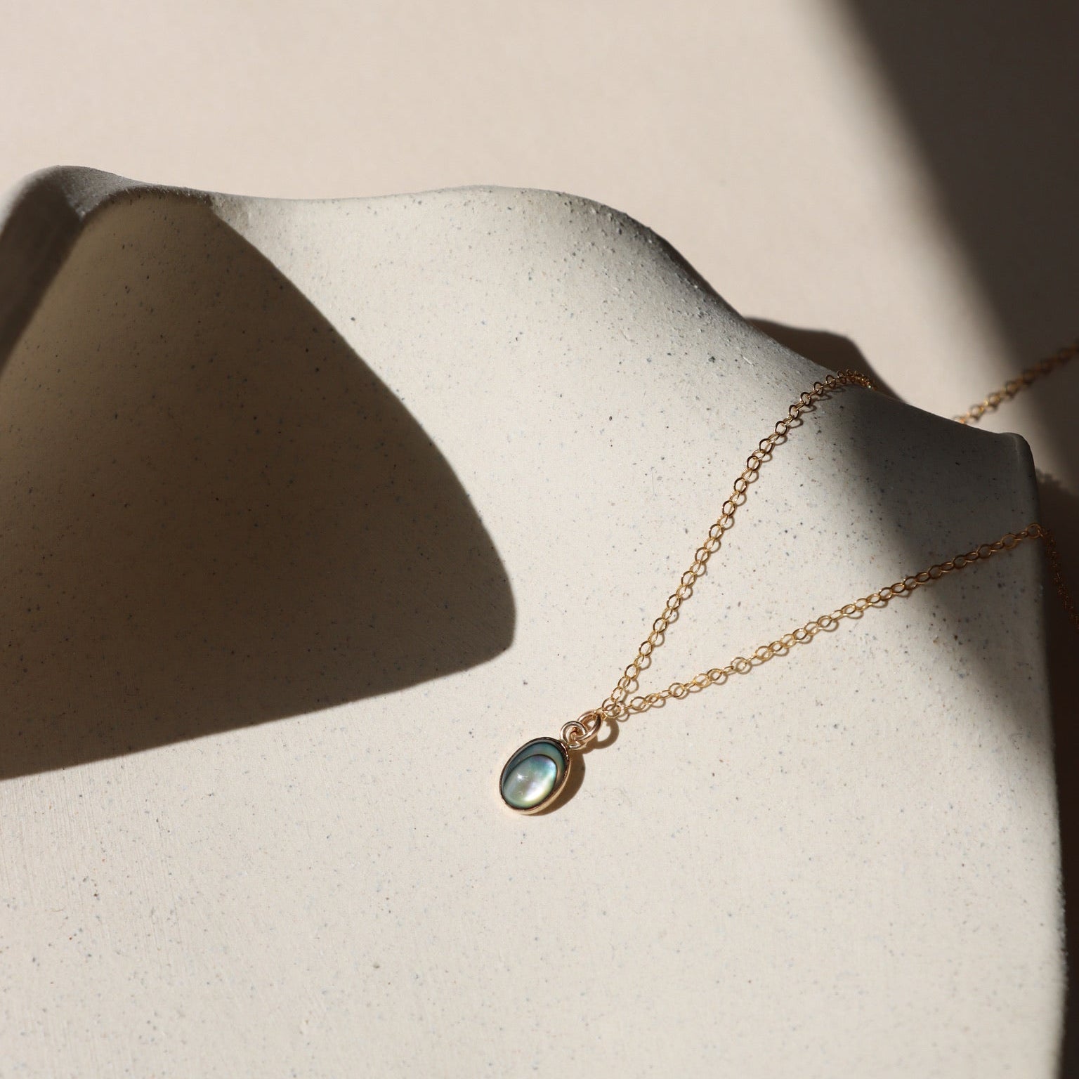 14k gold fill delicate chain necklace with 14k gold fill bezel pendant with an abalone stone on a light dish