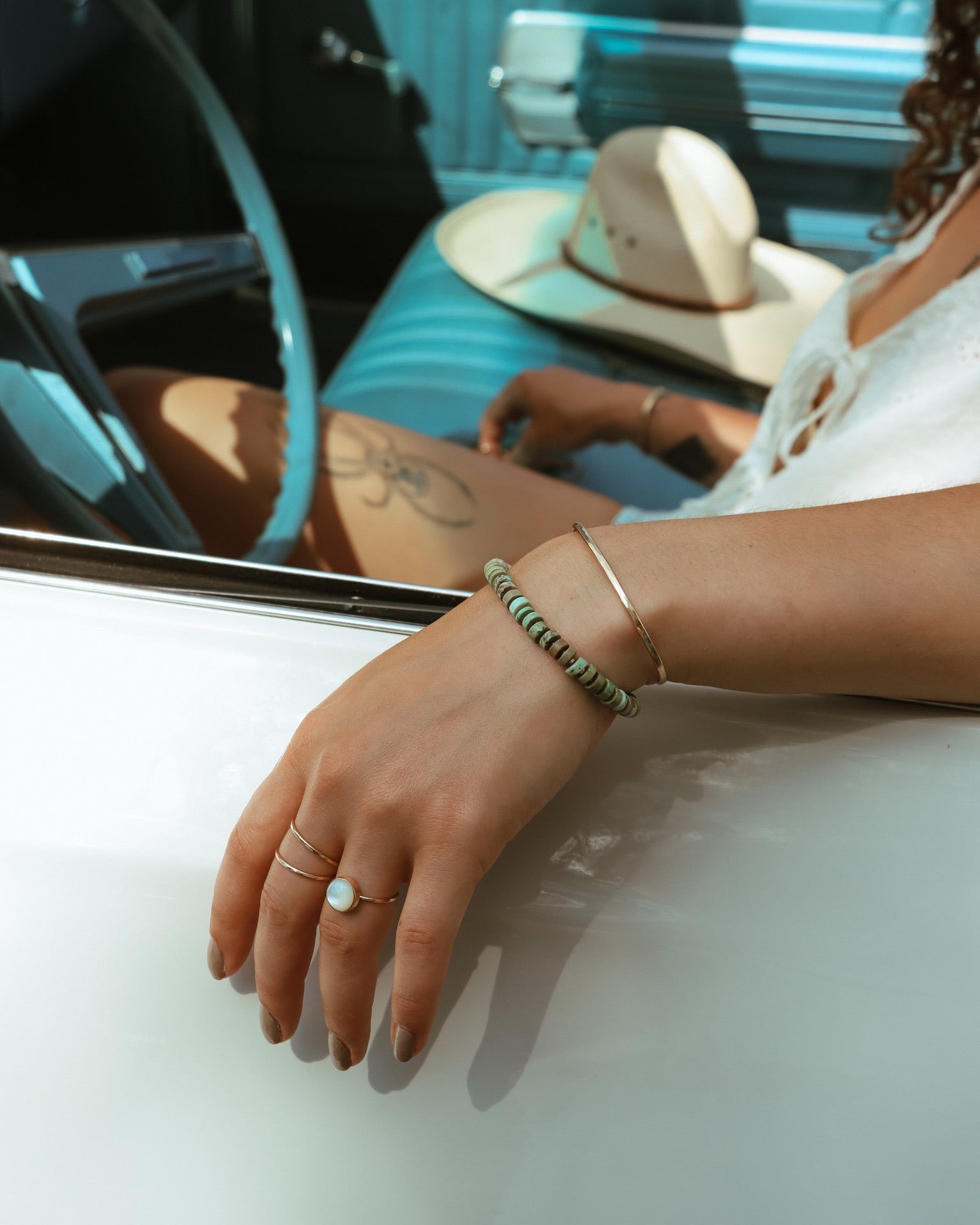 A hand resting on a vintage car door, displaying a stack of a beaded stone bracelet and a minimalist cuff alongside delicate stackable rings.