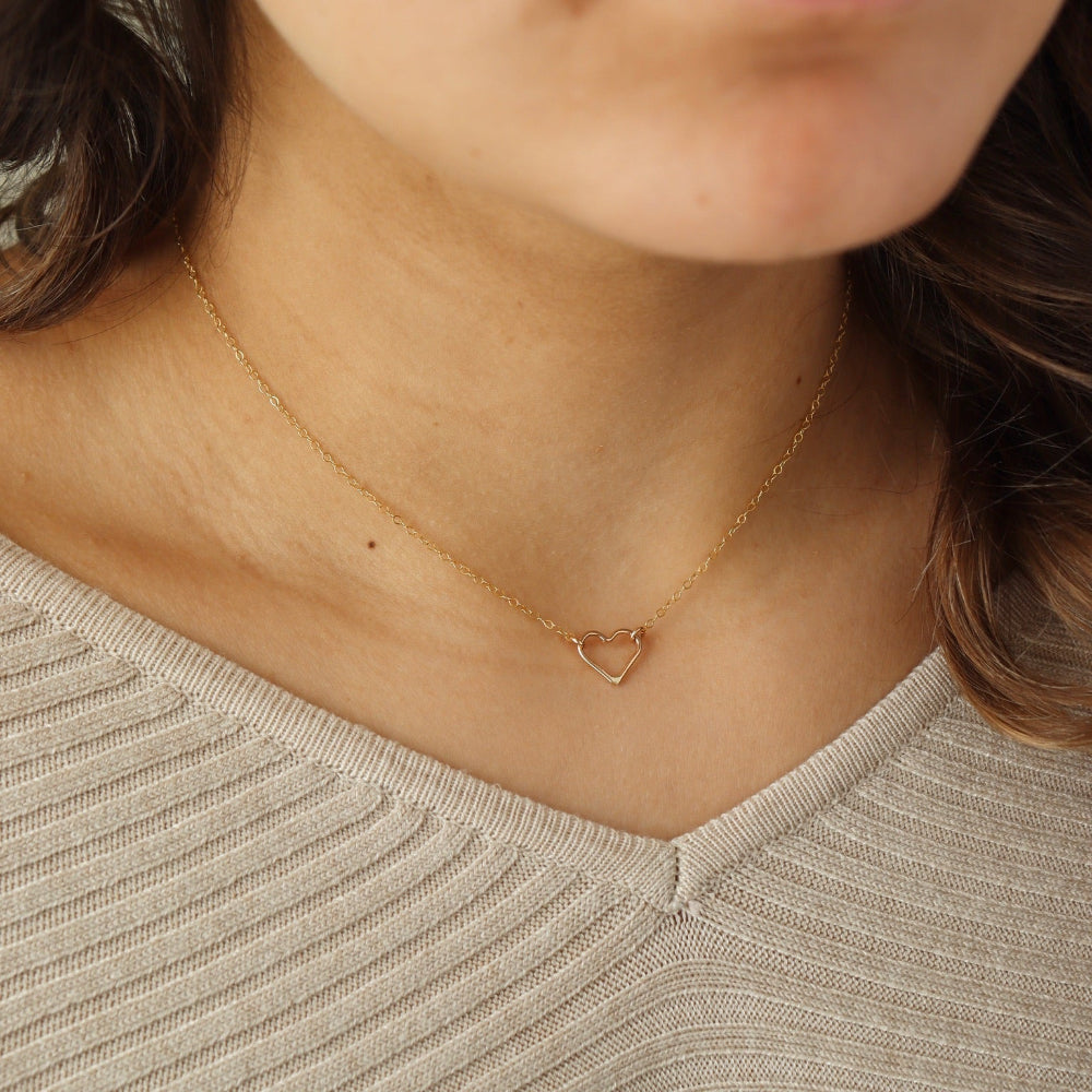 Model wearing a 14k gold fill necklace featuring a thin standard chain with an open heart pendant anchored at the end. 