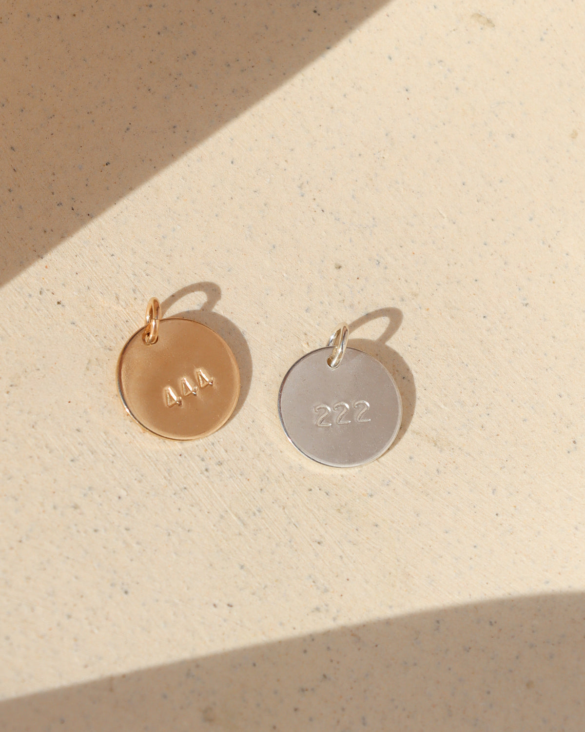 Two round angel number charms in gold and silver, stamped with 444 and 222, resting side by side on a textured cream surface in warm sunlight with soft shadows.