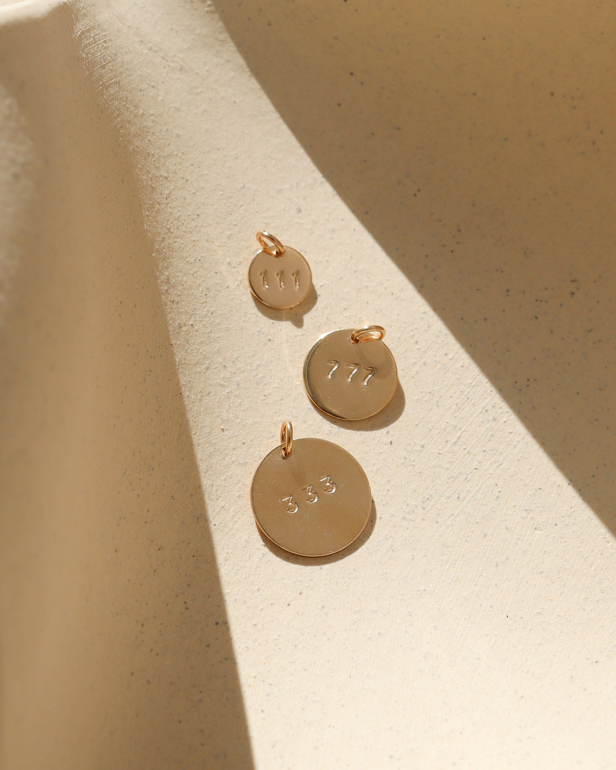 Three round gold angel number charms stamped with 111, 777, and 333 in graduated sizes, arranged on a textured cream surface with warm sunlight and soft shadows.