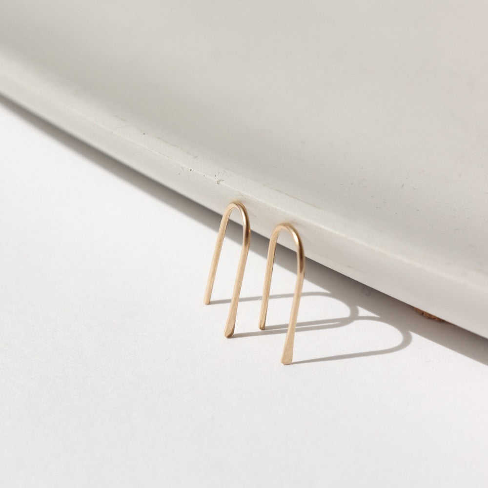 14k solid gold wire formed into arch shaped earrings with one hammered end, photographed on a white table leaning on a white dish