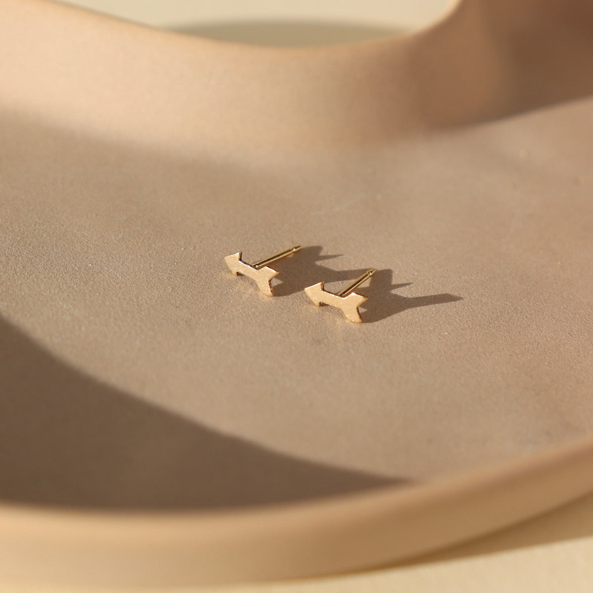 small gold arrow stud earrings on a sunlit ceramic dish