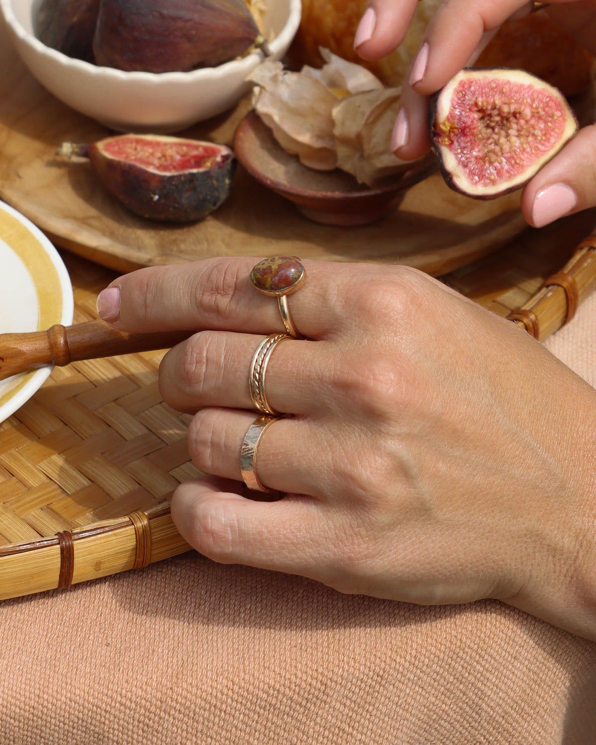 Hand wearing stacked gold rings and a colorful gemstone ring beside fresh figs in a warm, sunlit setting.