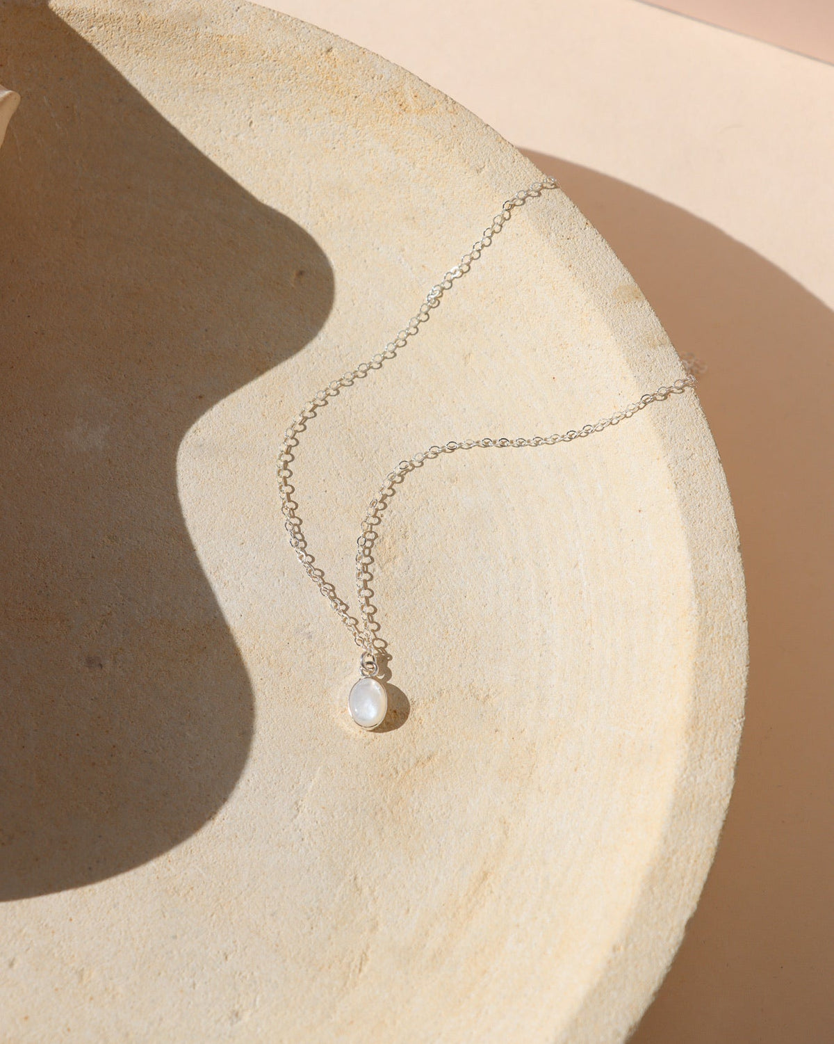 Delicate silver chain necklace featuring a small oval opal pendant in a silver bezel setting, displayed on a textured cream ceramic surface in warm natural sunlight with soft shadows.