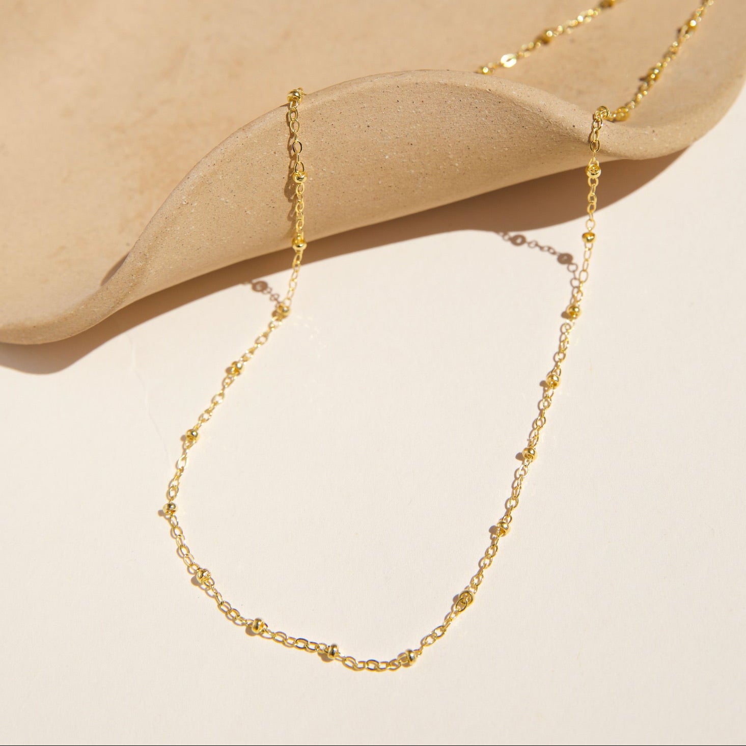 Top-down view of a fine gold satellite chain necklace with evenly spaced tiny bead accents, draped artistically over a minimalist wavy beige ceramic tray on a warm ivory surface
