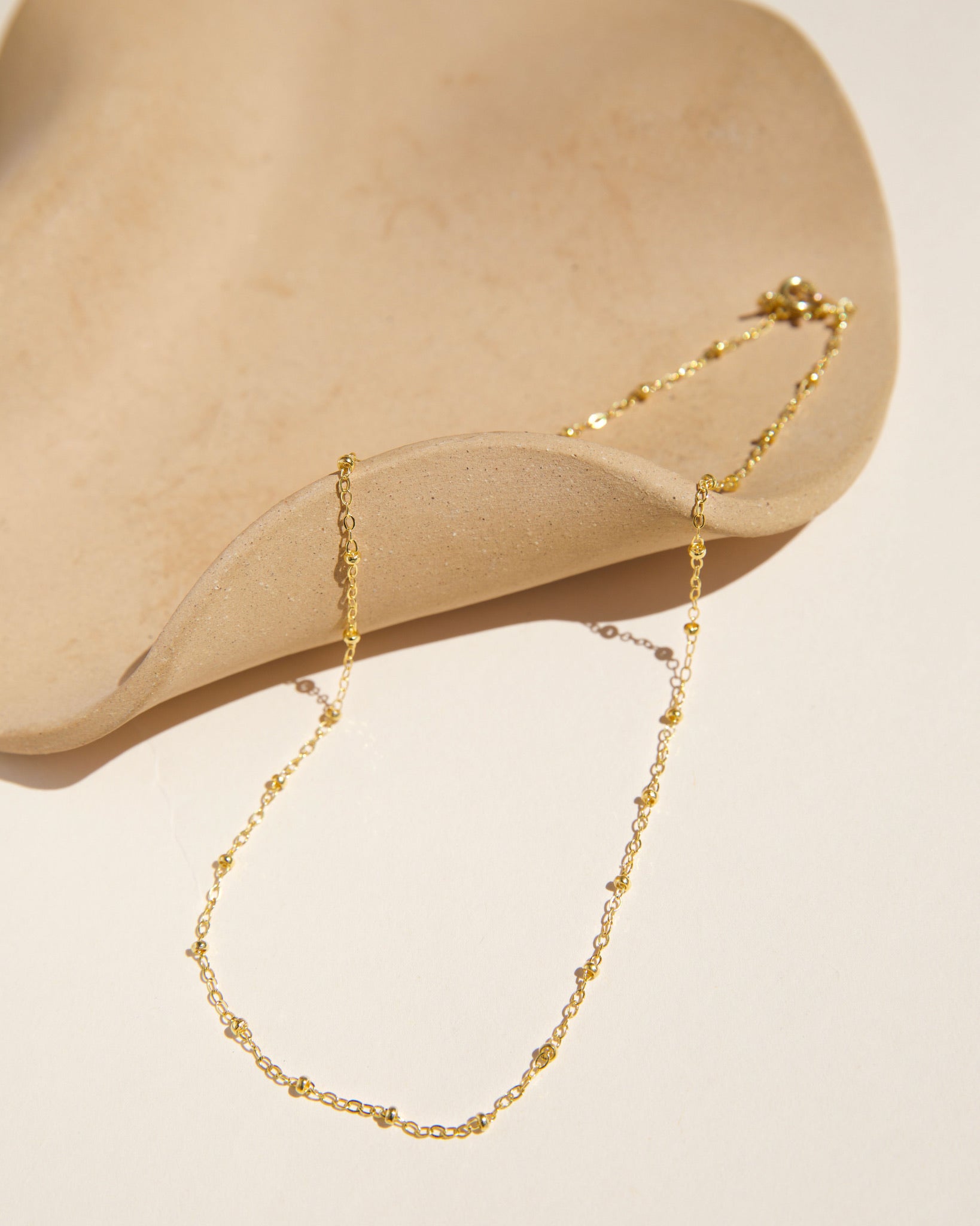 Top-down view of a fine gold satellite chain necklace with evenly spaced tiny bead accents, draped artistically over a minimalist wavy beige ceramic tray on a warm ivory surface