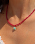 Close-up of a person wearing a bright pink beaded necklace with a small round gold pendant featuring a turquoise stone in a textured bezel, paired with a white ruffle-trim top in warm natural sunlight.