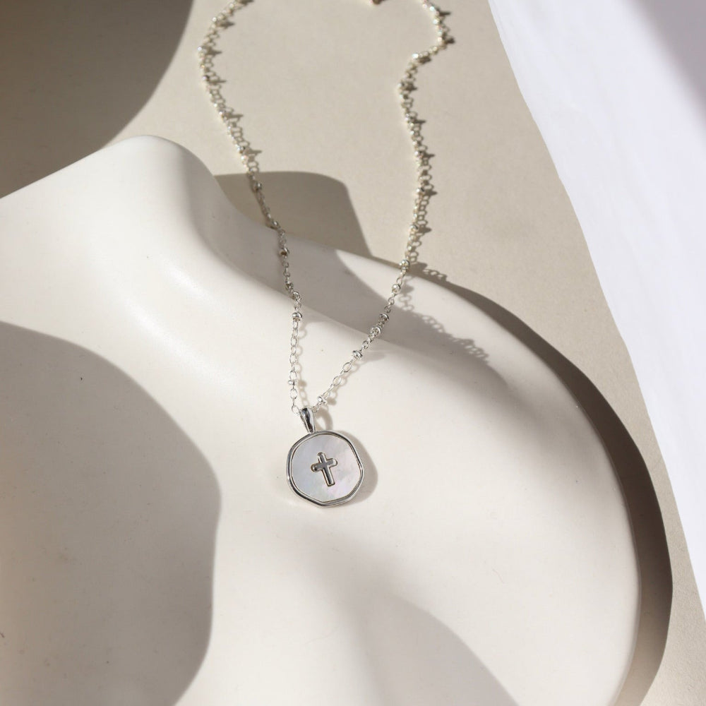 A small beaded sterling silver chain featuring a circle mother of pearl cross charm pendant. The necklace is displayed on a cream dish. 