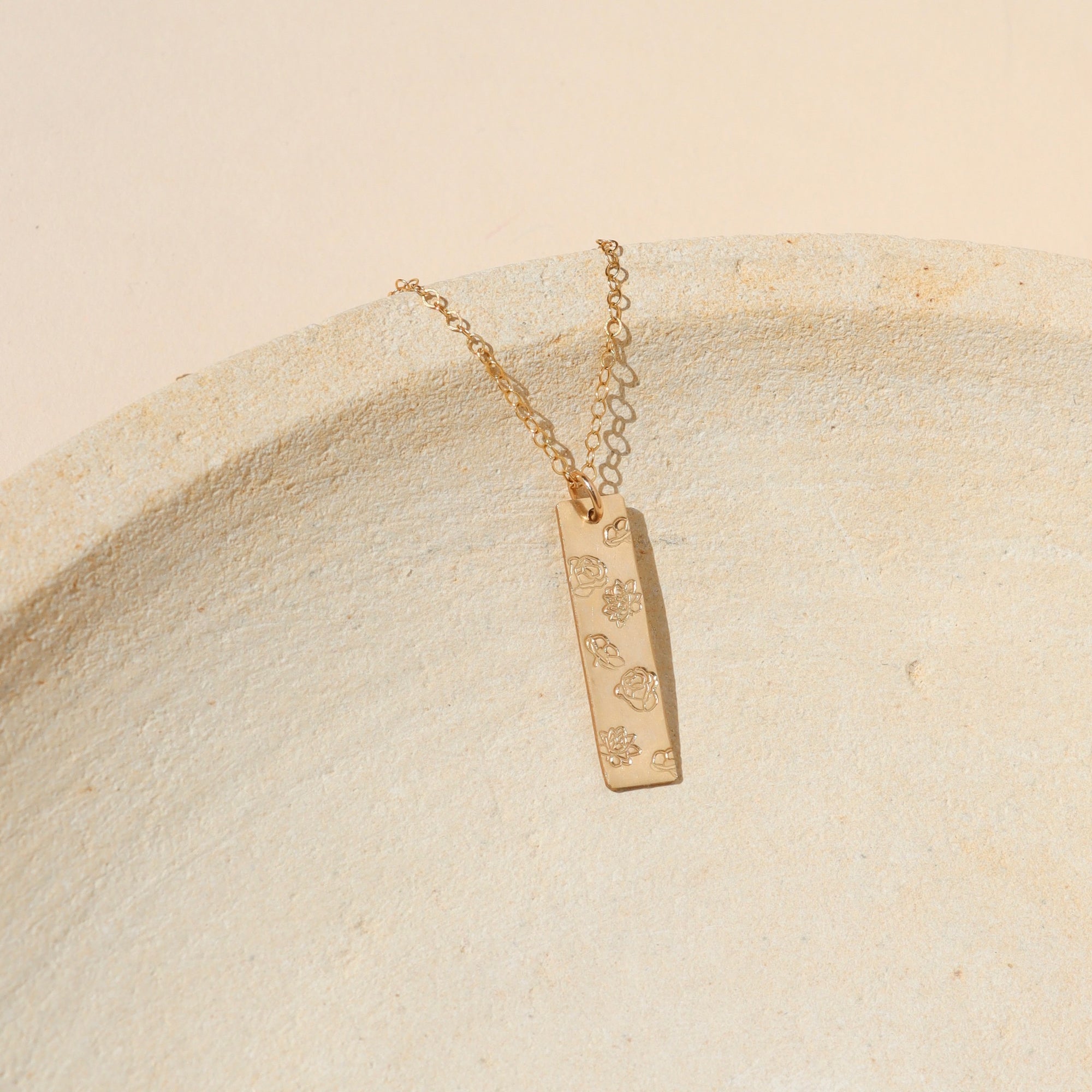 14k gold fill hand stamped bar pendant with flowers on a delicate chain on a light dish