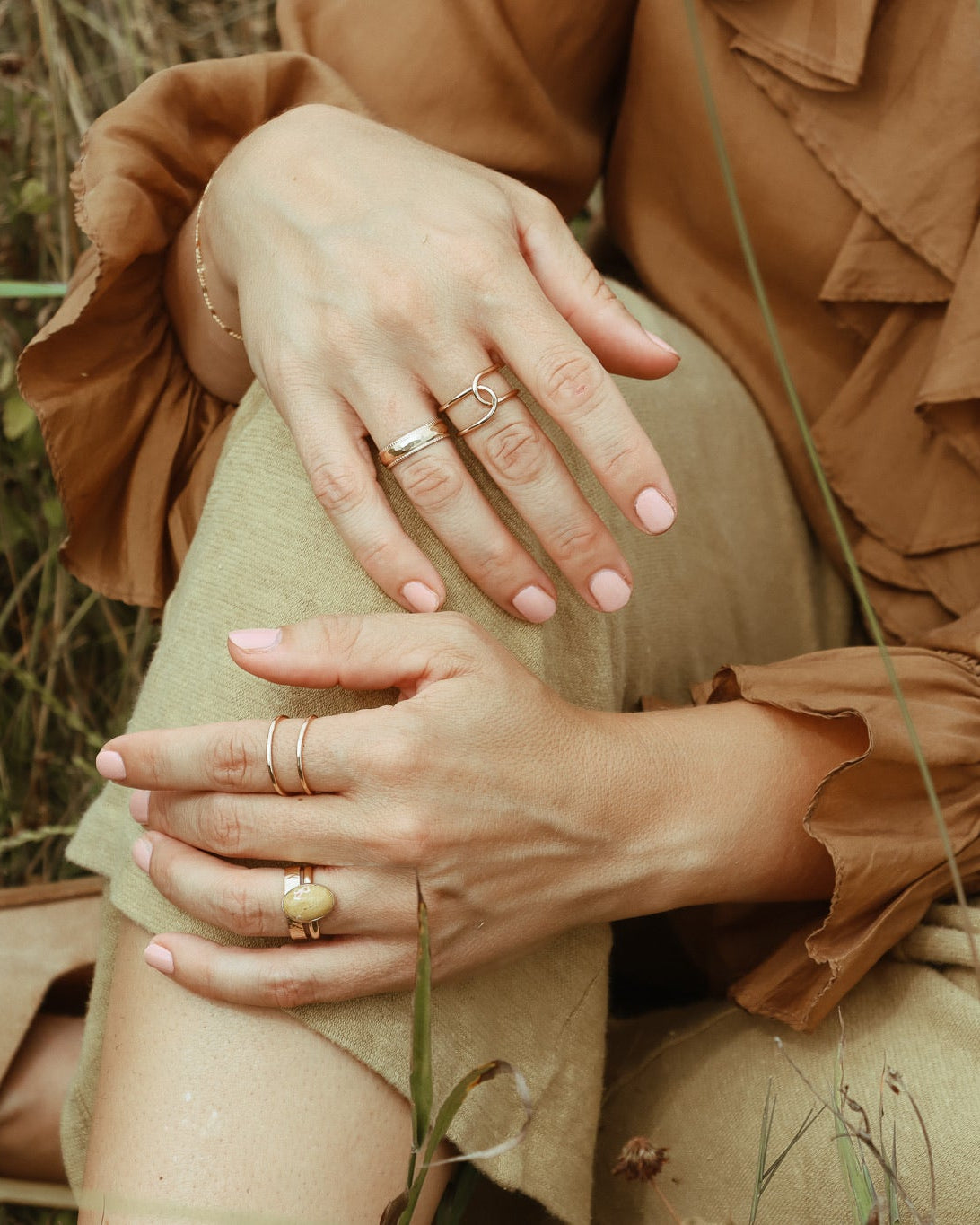 Close-up of layered gold rings and a gemstone ring styled with a warm brown blouse and neutral pants outdoors.
